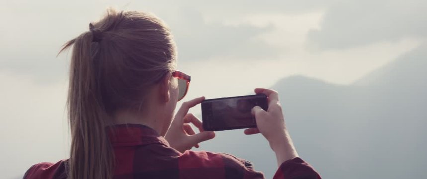 Blonde woman on a hike wear sunglasses take photo with her smartphone or cell phone or mobile hiking in autumn or fall cloud over mountain handheld over shoulder close up lens flare