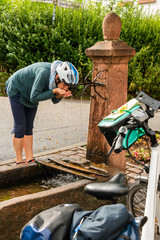 Fahrrad Fahrerin trinkt frisches Quellwasser am Dorfbrunnen, erholsame Unterbrechung einer Fahrrad Tour.