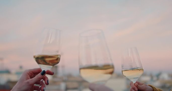 Hands cheering with glasses of white wine during the golden hour. With an old evening cityscape on their background. Slow Motion. 