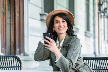 Young beautiful woman looking at smartphone while sitting at cafeteria. Happy university student using mobile phone. Businesswoman drinking coffee, smiling and using smartphone indoor.