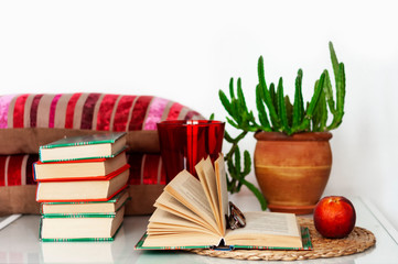 Cozy home interior decor: cup of coffee, stack of books, plants in pots, open book and red peach on a wicker stand, pillows on a white table. Distance home education.Quarantine concept of stay home.