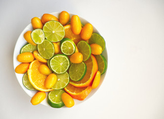 Sliced citrus fruit: lime, orange and kumquats on the white plate on white background. Minimal food concept. Flat lay, copy space, top view