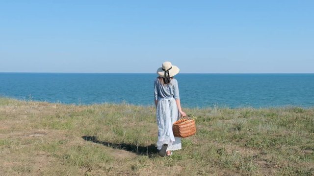 Young romantic tylish girl in vintage outfit walking on seaside with straw bag feeling inspired. Slow motion
