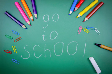 On a green chalkboard it is written to Go to school, next to it are crayons, pencils, colored paper clips, and a pen. Concept of the school, academic year. Beautiful, fun background for school.