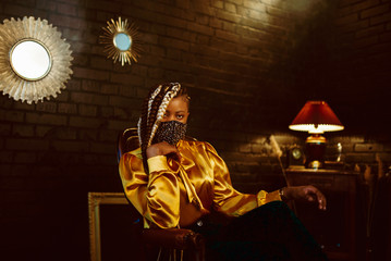 African American fashionable model wearing stylish dark green velvet protective face mask with golden rhinestones, yellow blouse, posing in dark vintage luxury interior. Copy, empty space for text