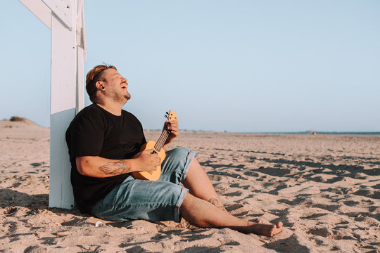 Hombre joven tocando el ukelele en la playa bajo la luz del sol durante la puesta de sol