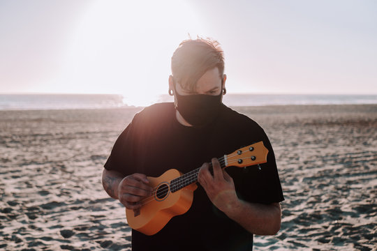 Hombre joven tocando el ukelele en la playa bajo la luz del sol durante la puesta de sol con una mascarilla, concepto de nueva normalidad