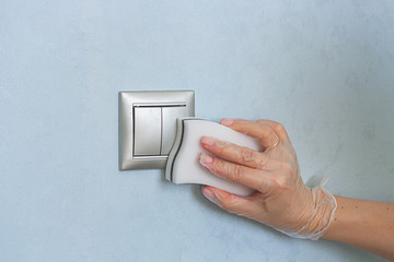 Hand in vinyl gloves is cleaning the light switch on blue wall by melamine sponge. Disinfecting surfaces in the house.