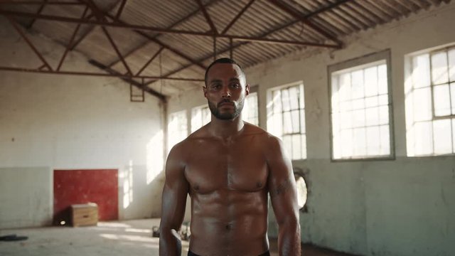 Fit young man standing shirtless at a gym inside abandoned warehouse. Determined sportsman in fitness studio looking at camera.
