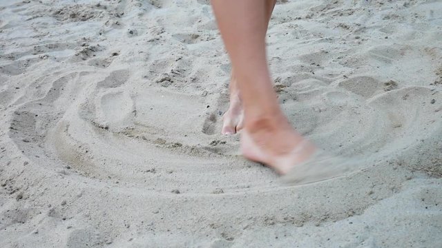 A woman draws circles in the sand with her toes and then brushes the sand towards the camera