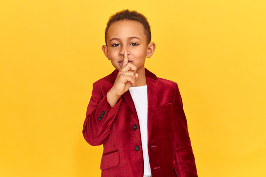 Cute dark skinned little boy making conspiracy gesture, keeping fore finger on his lips. Adorable African child saying shh, holding finger at mouth, asking to keep silence, trying to make no noise