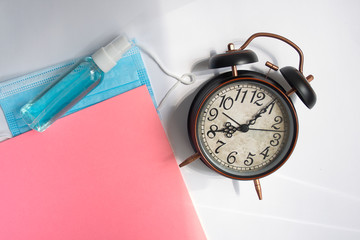 Alarm clock along with note blog and fountain pen, mask and disinfectant,pink paper,on gray background.