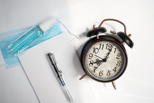 Alarm clock along with note blog and fountain pen, mask and disinfectant,white paper,on gray background.