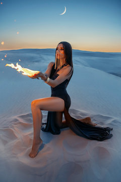 A mysterious woman sits on the sand and holds a fire in hands. Fantasy, art photo. Girl hides her face behind a golden veil. Lady in image of wizard. Desert night background. Creative dress with slits