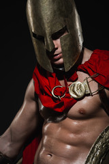 Young athletic sexy man in a costume of a Roman warrior in a red cloak and helmet on a black...