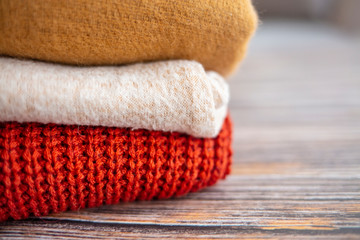 Bunch of knitted warm pastel color sweaters with different knitting patterns folded in stack on brown wooden table, grunged concrete wall background. Fall winter season knitwear. Close up, copy space