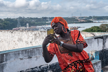 African woman with a mobile phone in hand and wearing traditional orange african dress. The place...