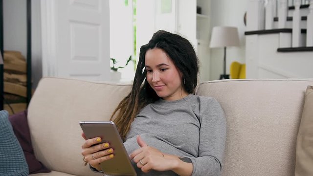 Pretty smiling happy young pregnant woman with dreadlocks relaxing on the soft couch at home and browsing photos on i-pad