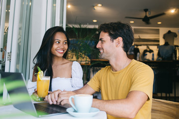 Smiling male and female multiracial friends having positive conversation about online project sitting in cafe with laptop computer, happy woman and man colleagues talking to each other on meeting