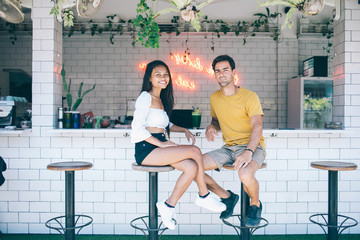 Cheerful multiracial male and female colleagues owner of cafeteria sitting on chairs looking at camera, portrait of trendy dressed 20s teen hipsters enjoying spending time together on free time