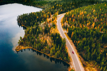 Aerial view of rural road with red car in yellow and orange autumn forest with blue lake