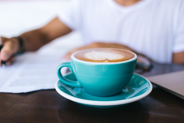 Selective focus on blue cup with caffeine beverage standing on working place with milky foram with coffee art on top, cropped image of hot coffee standing on blurred background with writing hand