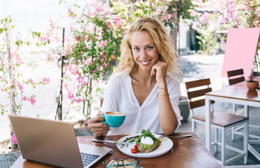 Obraz na płótnie Canvas Half length portrait of cheerful caucasian female digital nomad having break for brunch at table on cafe terrace,smiling woman freelancer sitting with laptop computer in restaurant eating healthy food