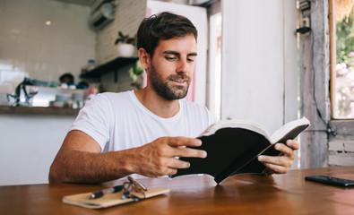 Handsome caucasian bearded male reader enjoying spending free time on hobby sitting in cafe interior, pensive young 20s hipster guy getting knowledge from literature holding book for education