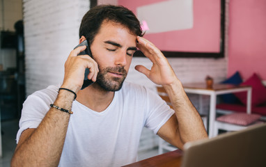 Sad caucasian male freelancer sitting in cafe interior on remote work making call to service operator for talking about trouble with internet connection, stress hipster guy holding hand feeling pain