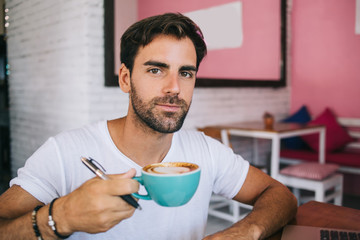 Portrait of handsome caucasian man writer holding coffee mug making break on job for enjoying aroma hot caffeine beverage, bearded male 20s looking at camera drinking cappuccino during works
