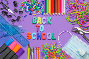 School stationery supplies with medical mask and hand sanitizer on purple background. Back to school and new normal concept. COVID-19 prevention.  Top view with copy space
