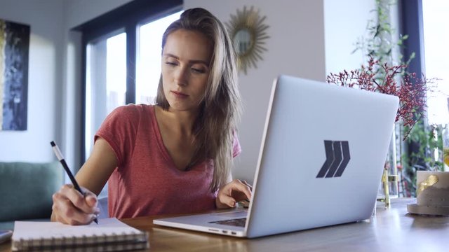 Young woman studying at home using laptop and taking notes