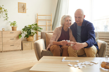 Warm toned portrait of happy senior couple holding domino pieces while playing board games at home,...