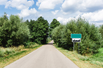 Asphalt road through the forest. Summer coniferous forest travel landscape. Long empty straight road and blue cloudy sky above. Empty copy space blank city name sign. Build up area road sign.