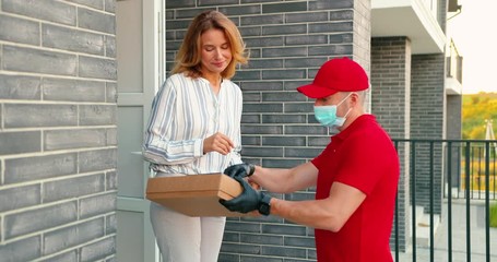 Caucasian male courier in red uniform and medical mask delivering carton parcel to female client at house and handing outdoor. Delivery man bringing boxes. Woman customer signing on tablet device.