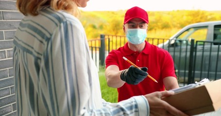 Caucasian male courier in medical mask delivering carton parcel to female client at house and handing outdoor. Delivery man bringing box to woman and she signing. Pandemic quarantine
