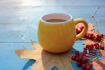 Yellow cup of coffee, red berries, and autumn leaves on blue wooden background.  Cozy autumn atmosphere. Copy space