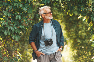 A mature man with a beard walks in the park with a camera. Active pastime in old age.