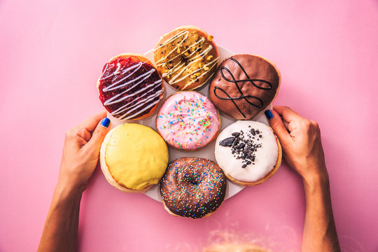 Close up of female hands holding dessert plate full of colorful chocolate donuts, on top of pink background.