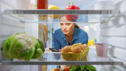 Young Woman Opens Fridge Door and Checks Recipe in Her Smartphone. Woman Preparing Healthy Meal....