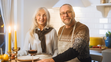 Happy Senior Couple in Love Have Romantic Evening, Smiling on Camera and Celebrating Anniversary. Elderly Have Romantic Evening with Wine, Festive Table in Cozy Kitchen Interior