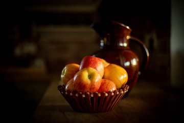 
apples in a basket, apples in a vase, a decanter behind a basket, a brown decanter, a decanter and a basket of apples on the table