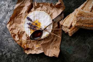 French cheese Camembert with spoon of honey, dried lavender stems on the paper  and freshly baked baguette over dark rustic background. Top view. Copy space