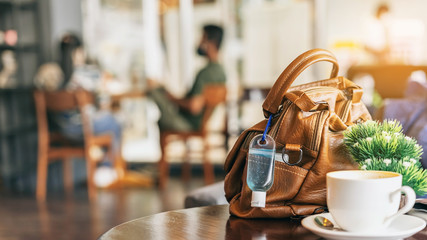 Mini alcohol gel bottle to kill Corona Virus(Covid-19) hang on a brown leather shoulder bag with hot coffee on table in cafe.New normal lifestyle.Health care concept.Selective focus on alcohol gel