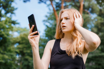 Naklejka premium Dissatisfied young hipster woman with blonde hair using mobile phone at the park. Emotion facial expression. Outdoors