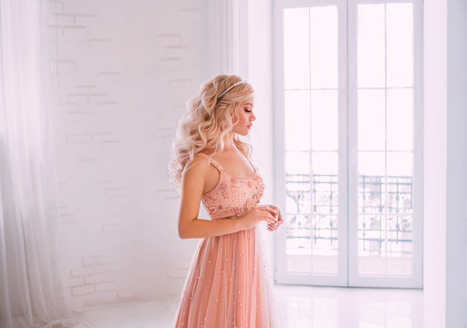 Portrait of a young beautiful woman. Closed eyes. Natural cosmetic. Pearl Tiara blond long wavy hair. Beige peach color luxurious dress embroidery. Backdrop white room interior, high classic window