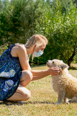 Healthcare and medicine concept. Blonde woman with the medicine mask on her face and blue dress pet densely curly beige dog in meadow during sunny summer day