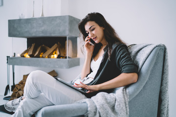 Fototapeta na wymiar Smiling caucasian female in casual wear sitting near fireplace in home interior talking on mobile phone and browsing on touchpad, positive woman calling to service for making delivery online