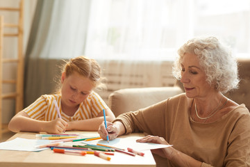 Warm toned portrait of smiling senior woman babysitting cute red haired girl and drawing together...