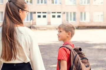 First day at school after holidays. Elder sister brings her younger brother to school. Boy carrying backpack, holding girl's hand, looking at her, walking to the entrance of school building.